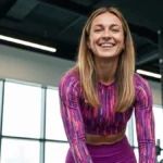 Anna Lewandowska uśmiecha się do aparatu, ubrana w elegancki strój sportowy, na tle nowoczesnego studia fitness